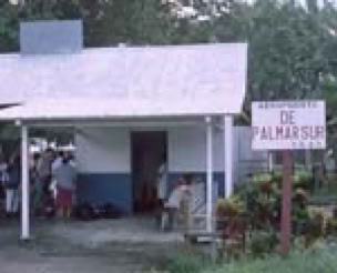 A small building with a white and blue facade, people gathered outside, and a sign reading "DE PALMIR SUR" next to it.